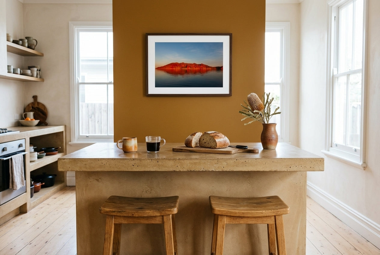Crimson Calm — Island Before Sunset, Lake Argyle, framed above a polished concrete breakfast counter on a deep ochre accent wall in a warm Australian kitchen — oak stools, dried banksia, dark wood frame. — Two Fat Bunnies.