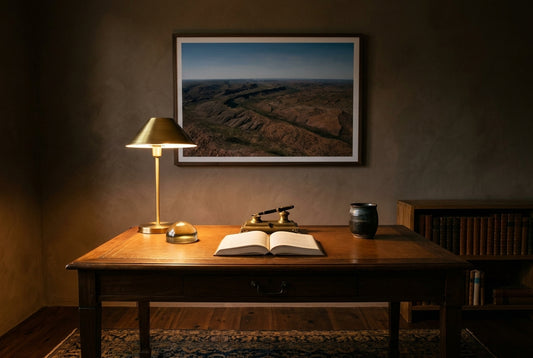 East Kimberley Ridgelines, framed above an aged walnut writing desk in a heritage home study — tan-leather inlay, brass-shaded reading lamp, leather-bound books, mushroom plaster walls, faded Persian rug, dark wood frame. — Two Fat Bunnies.