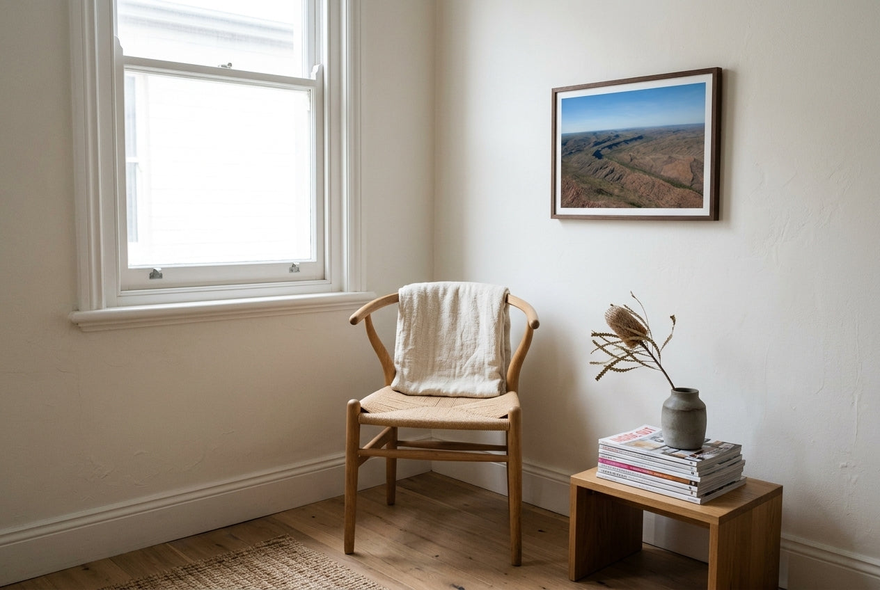 East Kimberley Ridgelines, framed beside an oak Wegner-style reading chair in a quiet reading nook — woven paper-cord seat, cream linen throw, small side table, dark wood frame. — Two Fat Bunnies.