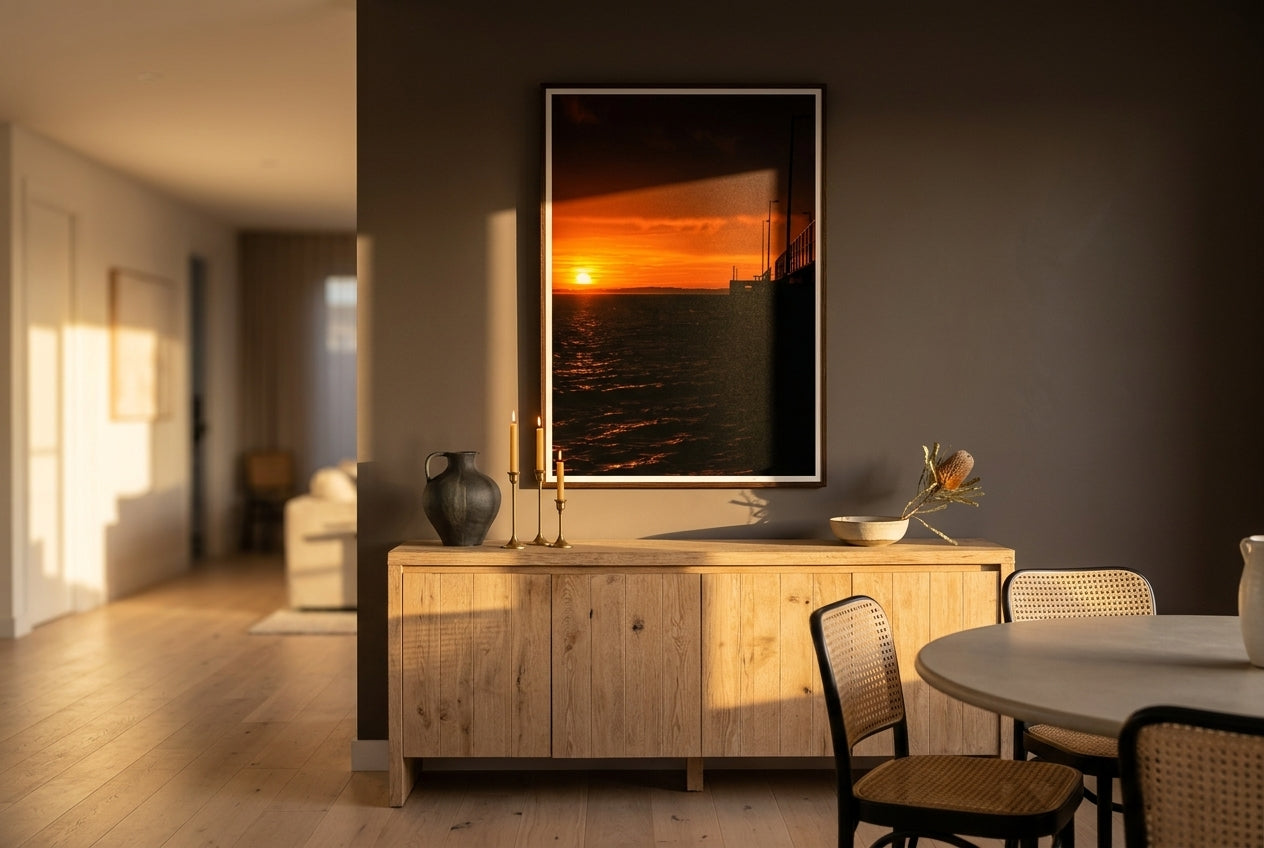 Golden Dawn at Flinders Pier, framed above a long raw-oak sideboard in a refined open-plan Australian living-dining space — sculptural ceramics, brass candleholders, dried banksia, dark wood frame. — Two Fat Bunnies.