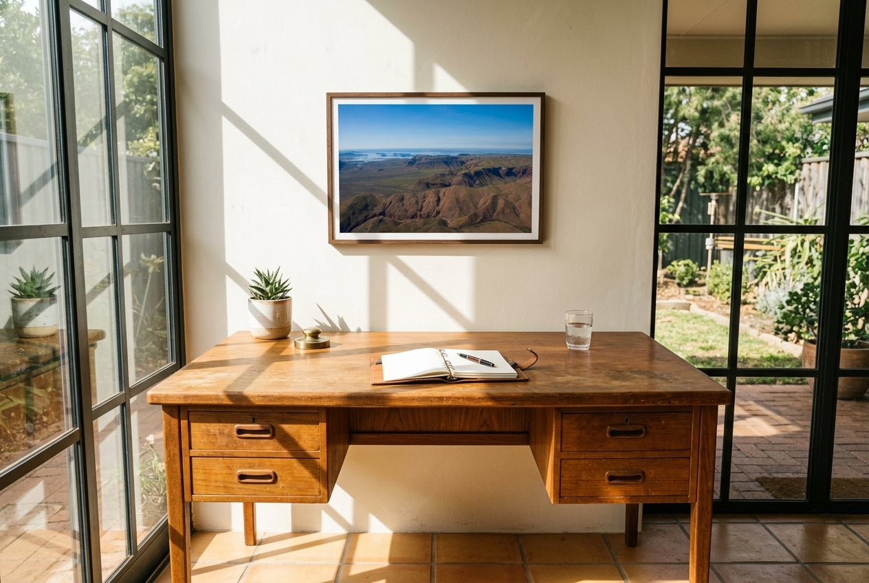 Lake Argyle From Above, framed above a vintage teak writing desk in a sun-drenched solar-garden room — terracotta tile, steel-framed glass garden door, succulent in clay pot, dark wood frame. — Two Fat Bunnies.