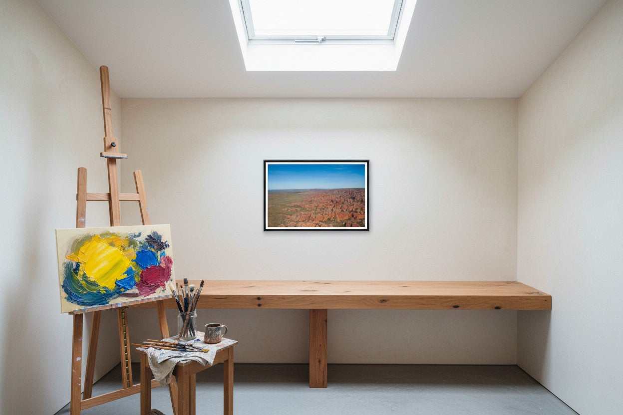 Over the Bungle Bungles, framed above the bench in an artist's home studio — raw-timber bench, timber easel, oil painting in progress, palette and brushes, north-facing skylight, dark wood frame. — Two Fat Bunnies.