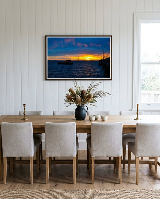 Sunrise Serenity at Flinders Pier, framed above a long oak dining table in a coastal dining room — woven linen runner, ceramic carafe, dried sea-grass in stoneware, woven rattan pendant overhead, dark wood frame. — Two Fat Bunnies.
