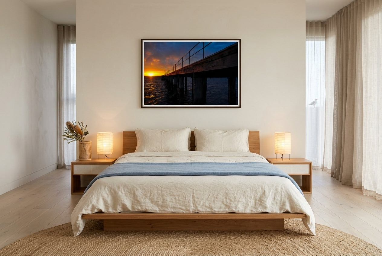 Dawn at Flinders Pier, framed above a low blackbutt platform bed in a coastal-Japandi master bedroom — cream linen, soft-blue wool throw, paper-shaded lamps, sheer linen drapes, dark wood frame. — Two Fat Bunnies.