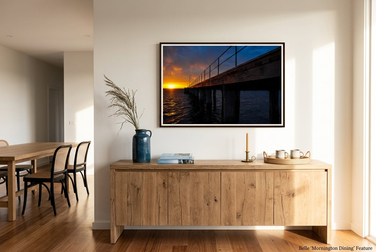 Dawn at Flinders Pier, framed above a long oak dining sideboard in a refined Mornington Peninsula dining room — coastal-blue ceramic vessel, dried sea-grass, photography books, brass candleholder, dark wood frame. — Two Fat Bunnies.