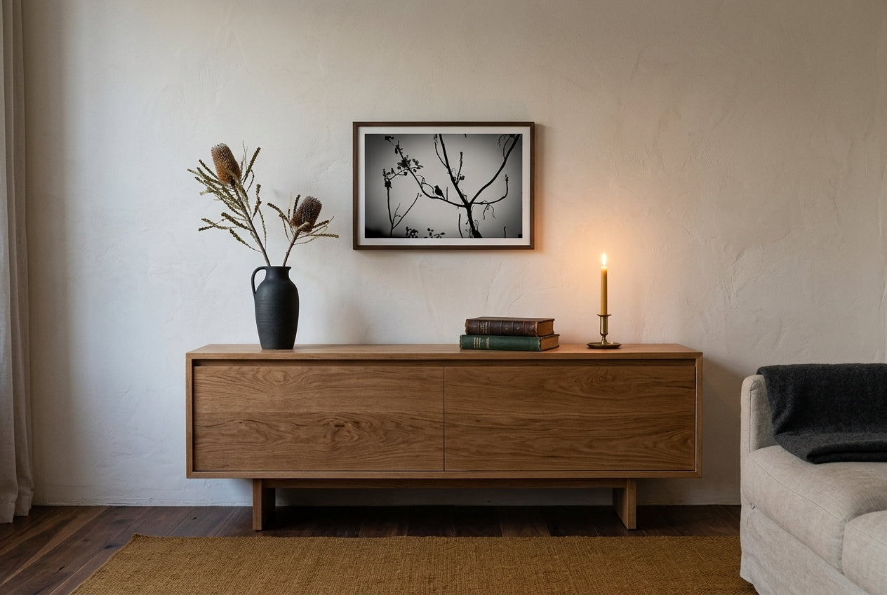 Dusk Solitude — El Questro Bird Silhouette, framed above a low oak sideboard in a contemplative Australian living room — charcoal ceramic with banksia, leather books, brass candle, dark wood frame. — Two Fat Bunnies.