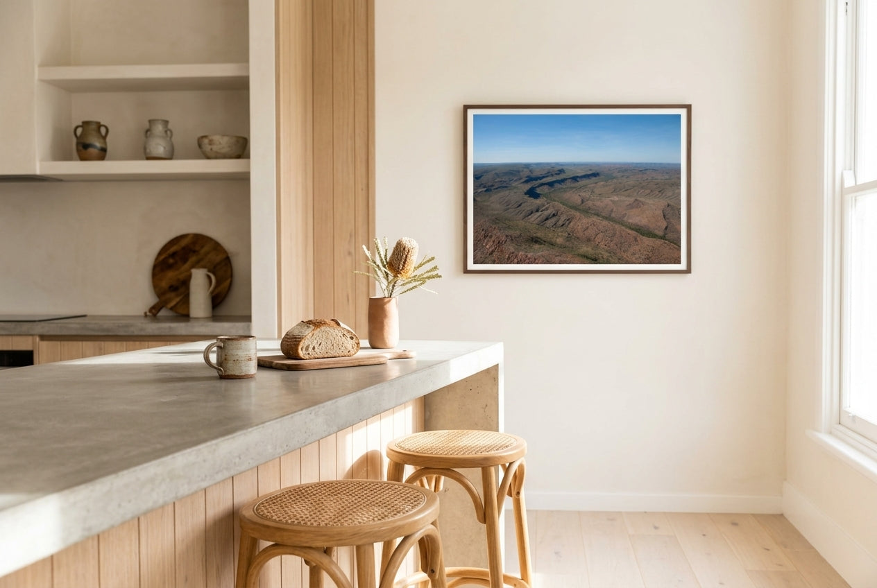 East Kimberley Ridgelines, framed above a polished concrete breakfast counter in a designer Australian kitchen — raw oak feature wall, oak-and-rattan stools, dried banksia, dark wood frame. — Two Fat Bunnies.