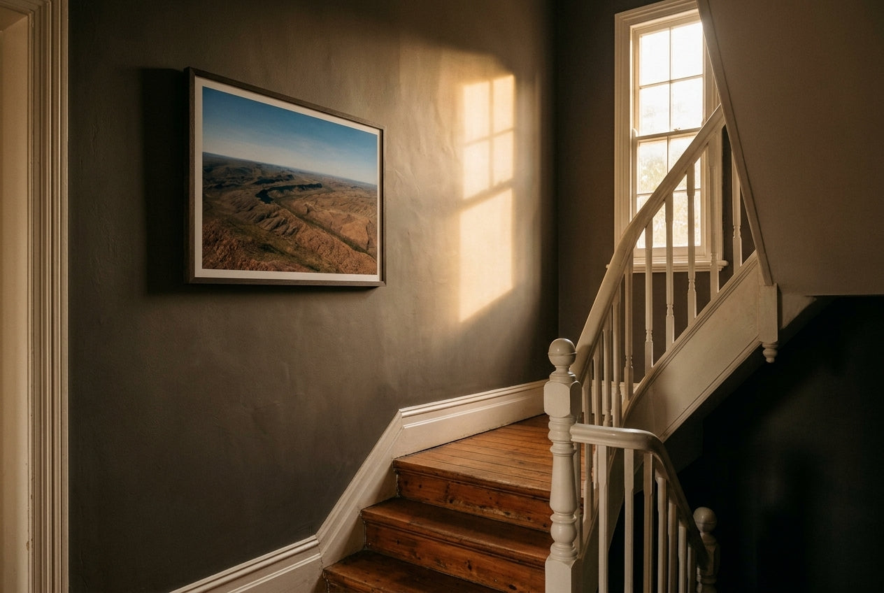 East Kimberley Ridgelines, framed as the single artwork on a heritage Australian stairwell landing — mushroom-charcoal plaster wall, high window casting directional light, dark wood frame. — Two Fat Bunnies.