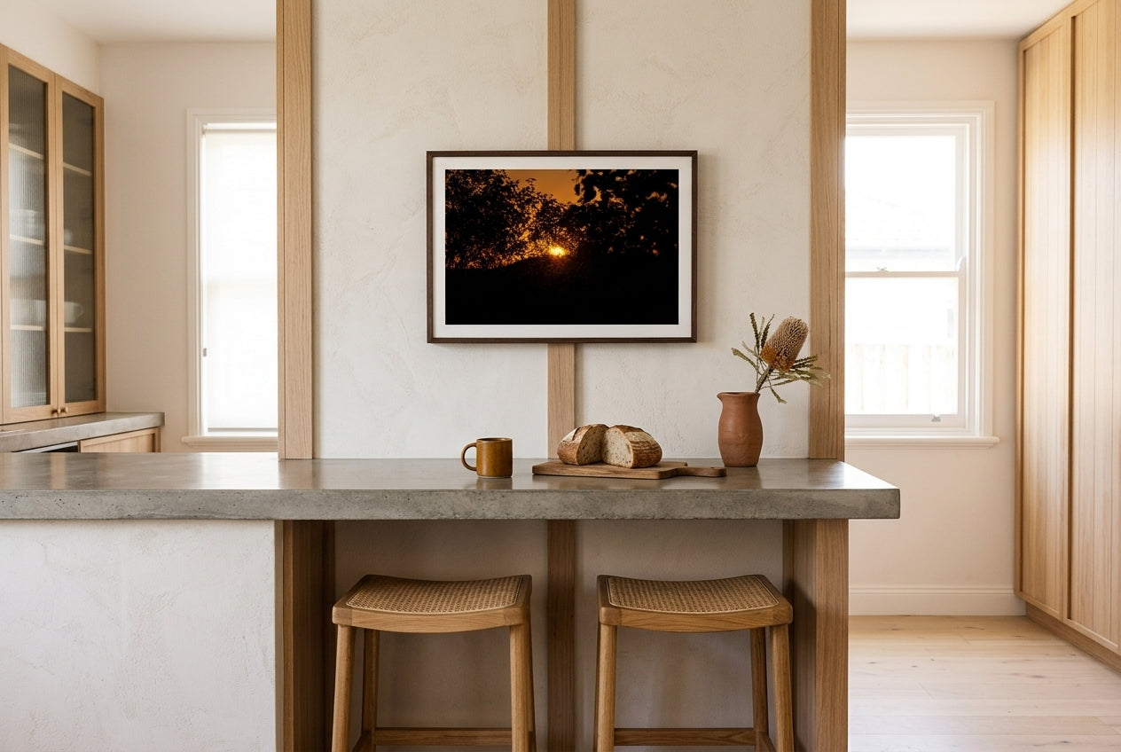 Ember Set at Buddy's Lookout, El Questro, framed above a polished concrete breakfast counter in a designer Australian kitchen — oak-and-rattan stools, dried banksia, ceramic vessel, dark wood frame. — Two Fat Bunnies.