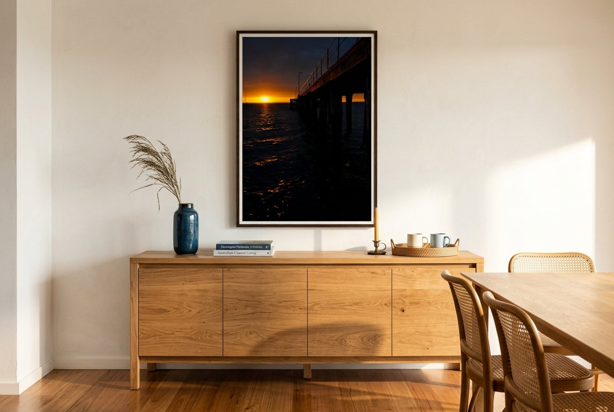 Flinders Sunrise Glow, framed above a long oak dining sideboard in a refined Mornington Peninsula dining room — coastal-blue ceramic, dried sea-grass, photography books, brass candle, polished blackbutt floors, dark wood frame. — Two Fat Bunnies.