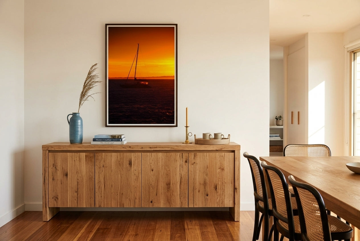 Golden Dawn at Flinders – Sunrise Boat, framed above a long oak dining sideboard in a refined Mornington Peninsula dining room — coastal-blue ceramic, dried sea-grass, photography books, brass candle, polished blackbutt floors, dark wood frame. — Two Fat Bunnies.