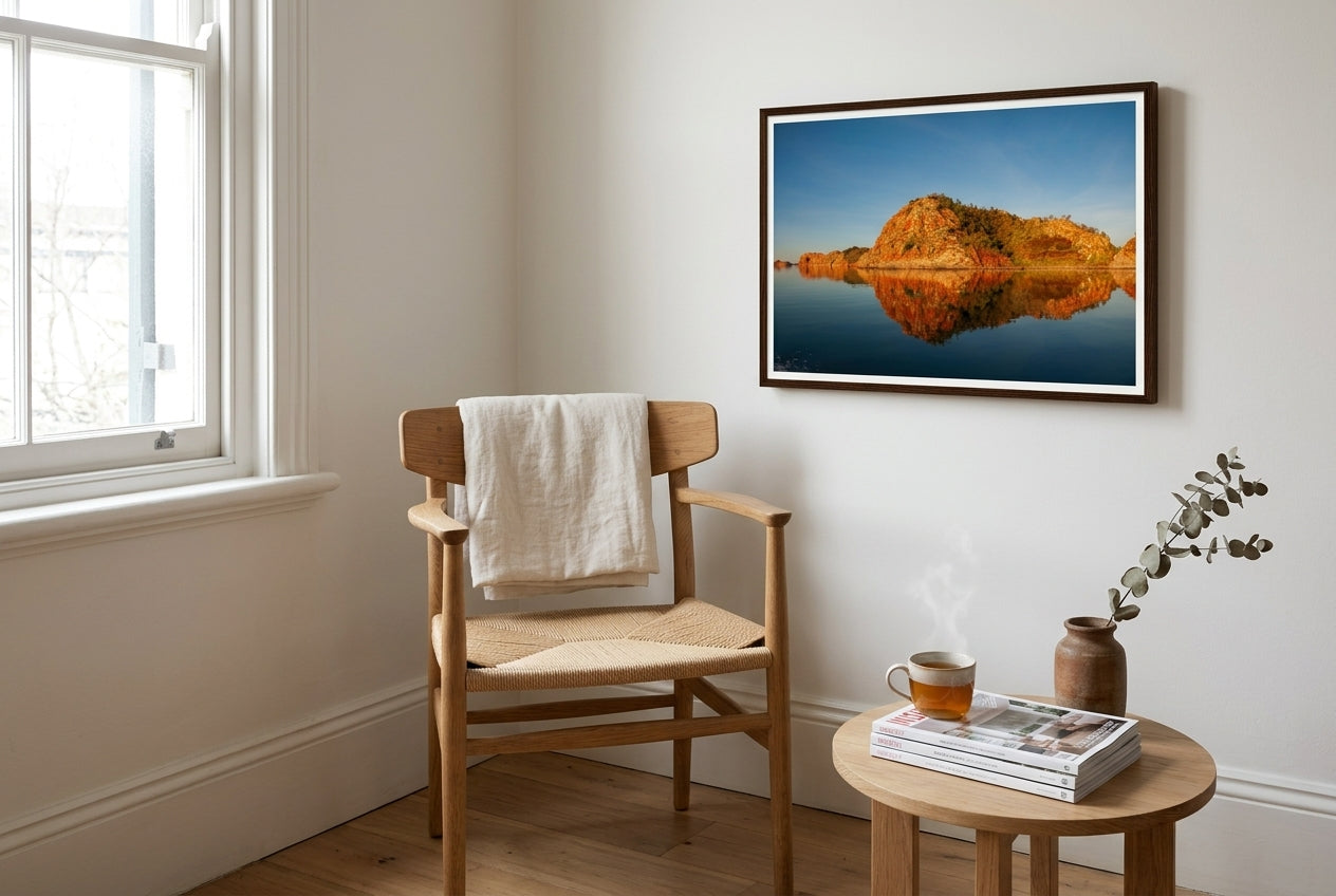 Golden Hour Mirror — Island Reflections, Lake Argyle, framed beside an oak Wegner-style reading chair in a Sydney apartment — cream linen throw, side table, sash window, dark wood frame. — Two Fat Bunnies.