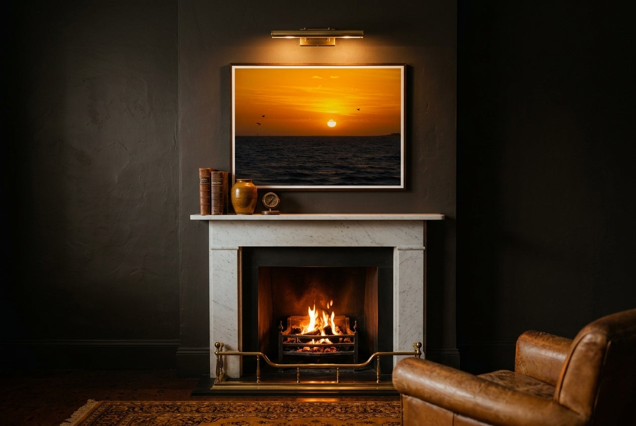 Golden Sunset Over Port Phillip Bay, framed above a charcoal snug mantelpiece — brass fender, low ember fire, leather club chair, dark wood frame. — Two Fat Bunnies.