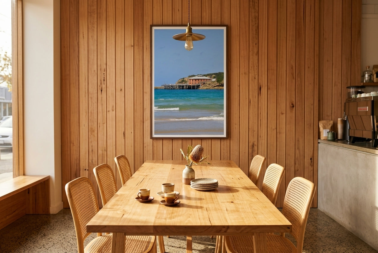 Heritage Tranquillity: Tathra Wharf, framed above a raw timber dining table — woven cane chairs, single brass pendant, espresso machine, dark wood frame. — Two Fat Bunnies.