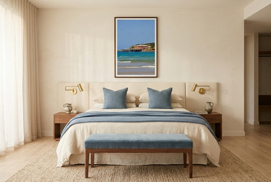 Heritage Tranquillity: Tathra Wharf, framed above an upholstered linen headboard in a boutique hotel suite bedroom — layered cream and dusty-blue bedding, polished brass fixtures, ottoman at foot of bed, dark wood frame. — Two Fat Bunnies.