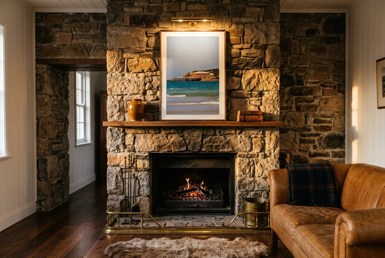 Heritage Tranquillity: Tathra Wharf, framed above a stone fireplace in a country Australian homestead living room — cast-iron tools, tartan throw, leather settee, sheepskin rug, dark wood frame. — Two Fat Bunnies.