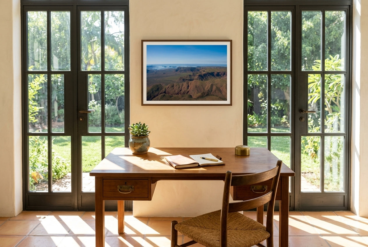 Lake Argyle From Above, framed above a vintage teak writing desk in a solar-garden room — terracotta tile, steel-framed glass garden door, succulent in clay pot, dark wood frame. — Two Fat Bunnies.