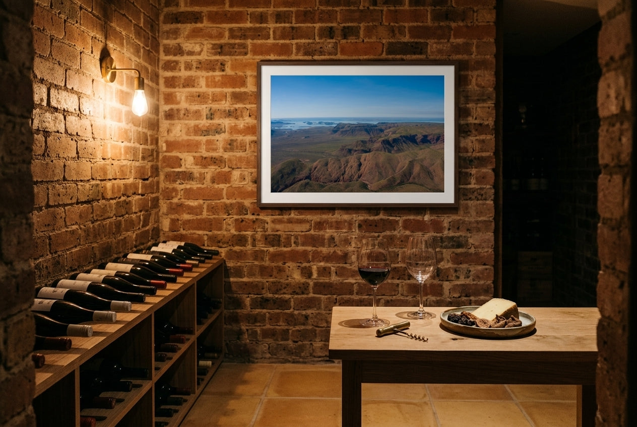 Lake Argyle From Above, framed in a small wine cellar tasting nook — corked bottles in timber racks, raw oak tasting table, crystal stemmed glasses, exposed brick wall, dark wood frame. — Two Fat Bunnies.