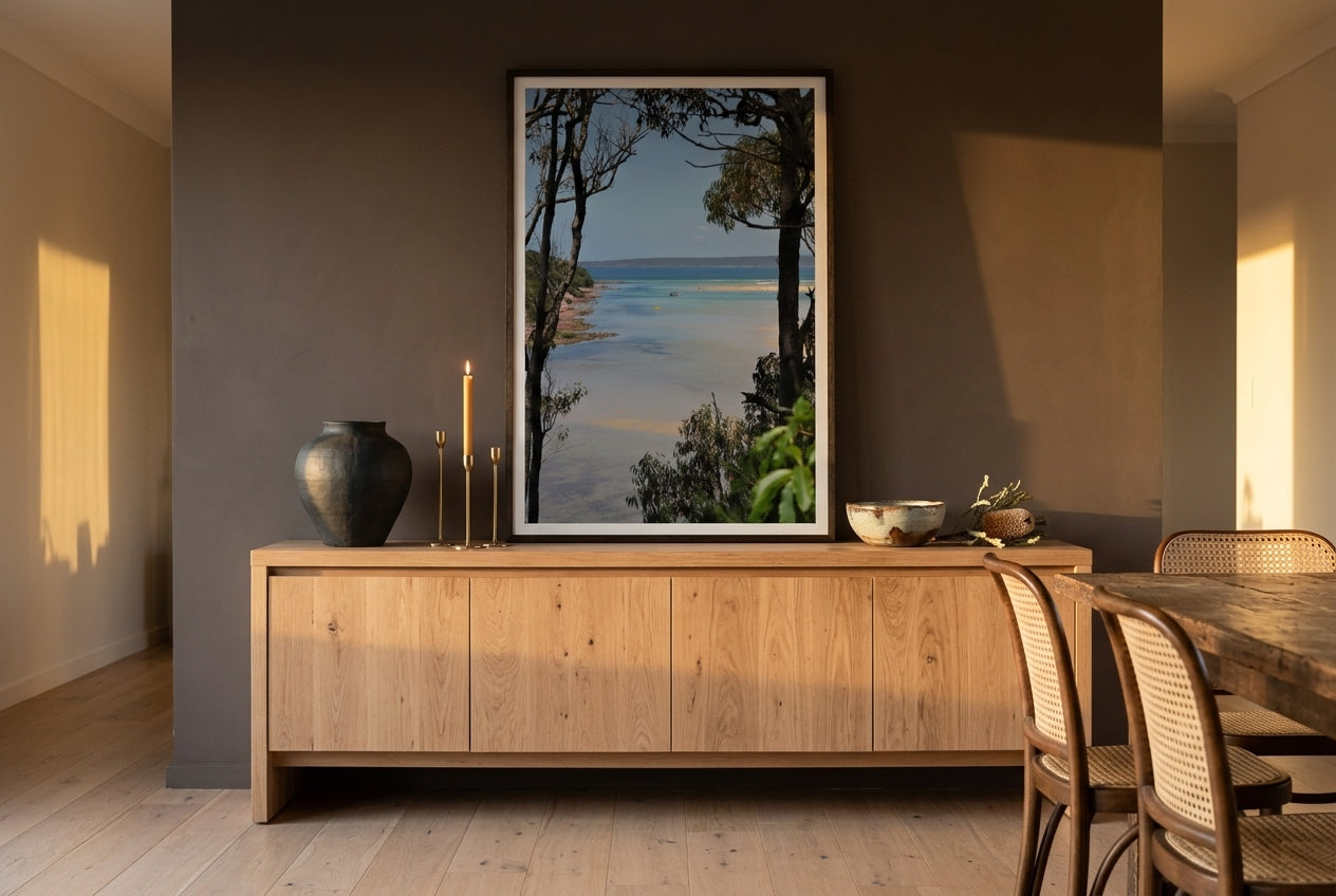 Merimbula Bliss, framed above a long raw-oak sideboard in a refined open-plan Australian living-dining space — sculptural ceramics, brass candleholders, dried banksia, dark wood frame. — Two Fat Bunnies.