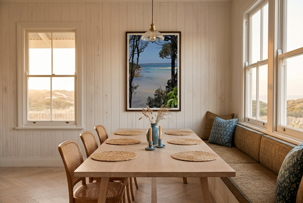 Merimbula Bliss, framed above a long oak dining table in a coastal weatherboard cottage — whitewashed timber walls, banquette with sea-grass cushions, ceramic blue vase, sash windows, dark wood frame. — Two Fat Bunnies.