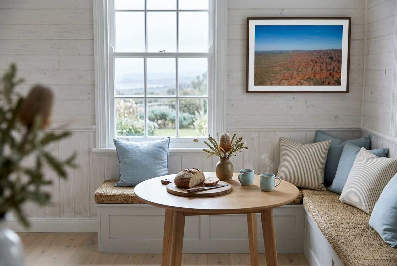 Over the Bungle Bungles, framed above the bench in a coastal cottage breakfast nook — whitewashed timber walls, sea-grass bench cushion, pale blue ceramic mugs, dried banksia in clay vessel, dark wood frame. — Two Fat Bunnies.