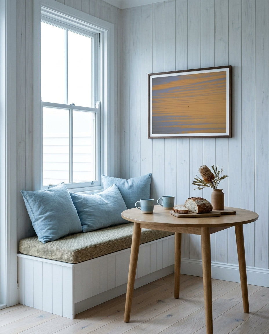 Solitude at Merimbula, framed in a coastal cottage breakfast nook — banquette seating, painted timber walls, ceramic mug, dark wood frame. — Two Fat Bunnies.