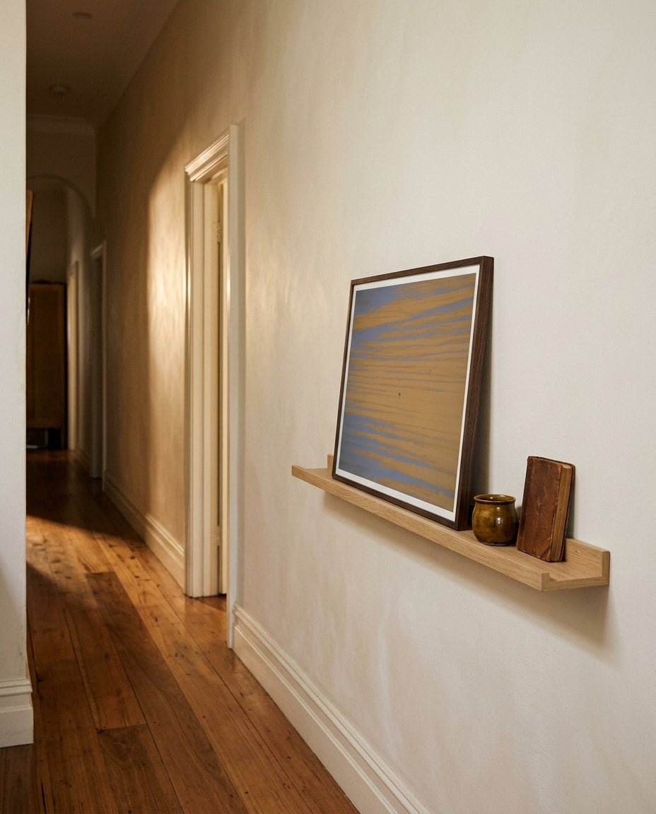 Solitude at Merimbula, framed on a hallway picture ledge — pale plaster wall, sculptural vessel, runner rug, dark wood frame. — Two Fat Bunnies.
