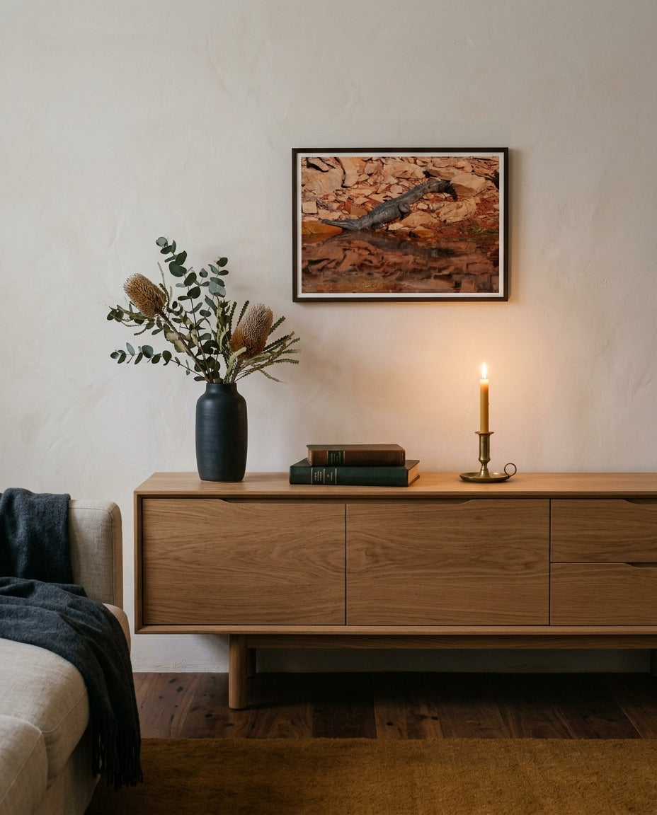Sunlit Stillness — Freshwater Crocodile, Lake Argyle, framed above a low oak sideboard in an Australian living room — charcoal ceramic with banksia, leather books, brass candle, dark wood frame. — Two Fat Bunnies.