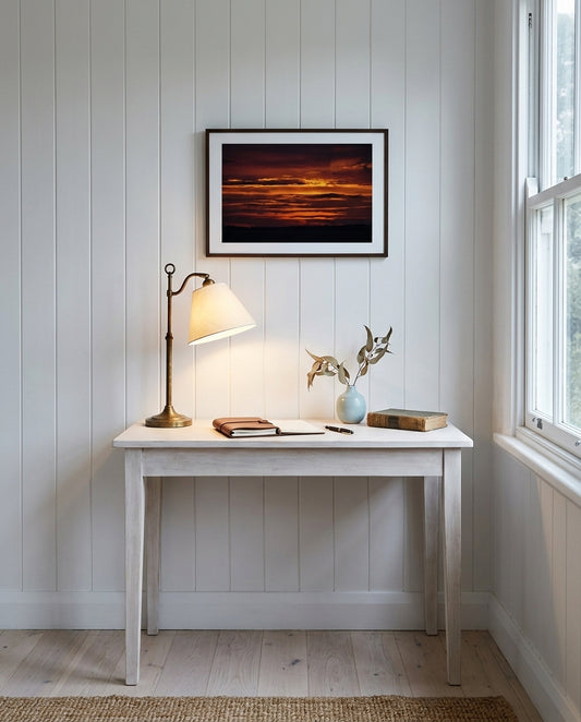 Sunrise over Bass Strait: Dawn at Flinders, framed above a slim oak writing desk in a coastal writing alcove — woven cane chair, ceramic mug, brass desk lamp, stack of paperbacks, dark wood frame. — Two Fat Bunnies.
