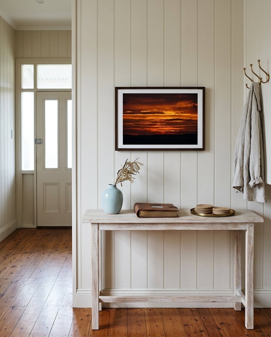 Sunrise over Bass Strait: Dawn at Flinders, framed above a slim console in a weatherboard hallway — limewash walls, woven cane basket, ceramic vessel with eucalyptus, brass key dish, dark wood frame. — Two Fat Bunnies.