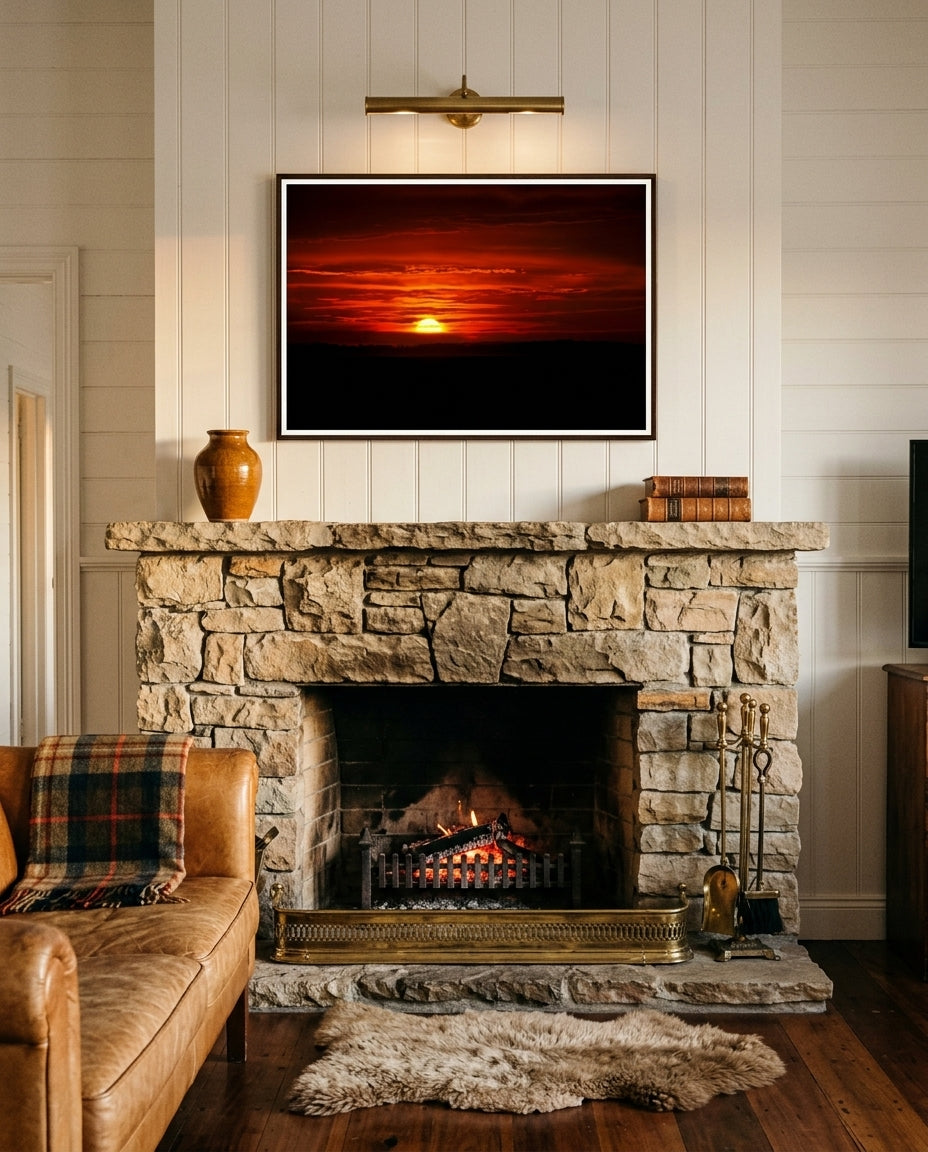 Sunrise Over Bass Strait – Flinders, framed above a stone fireplace in a country homestead living room — wool blanket on a leather wingback, brass fire tools, stacked firewood, woollen rug, dark wood frame. — Two Fat Bunnies.