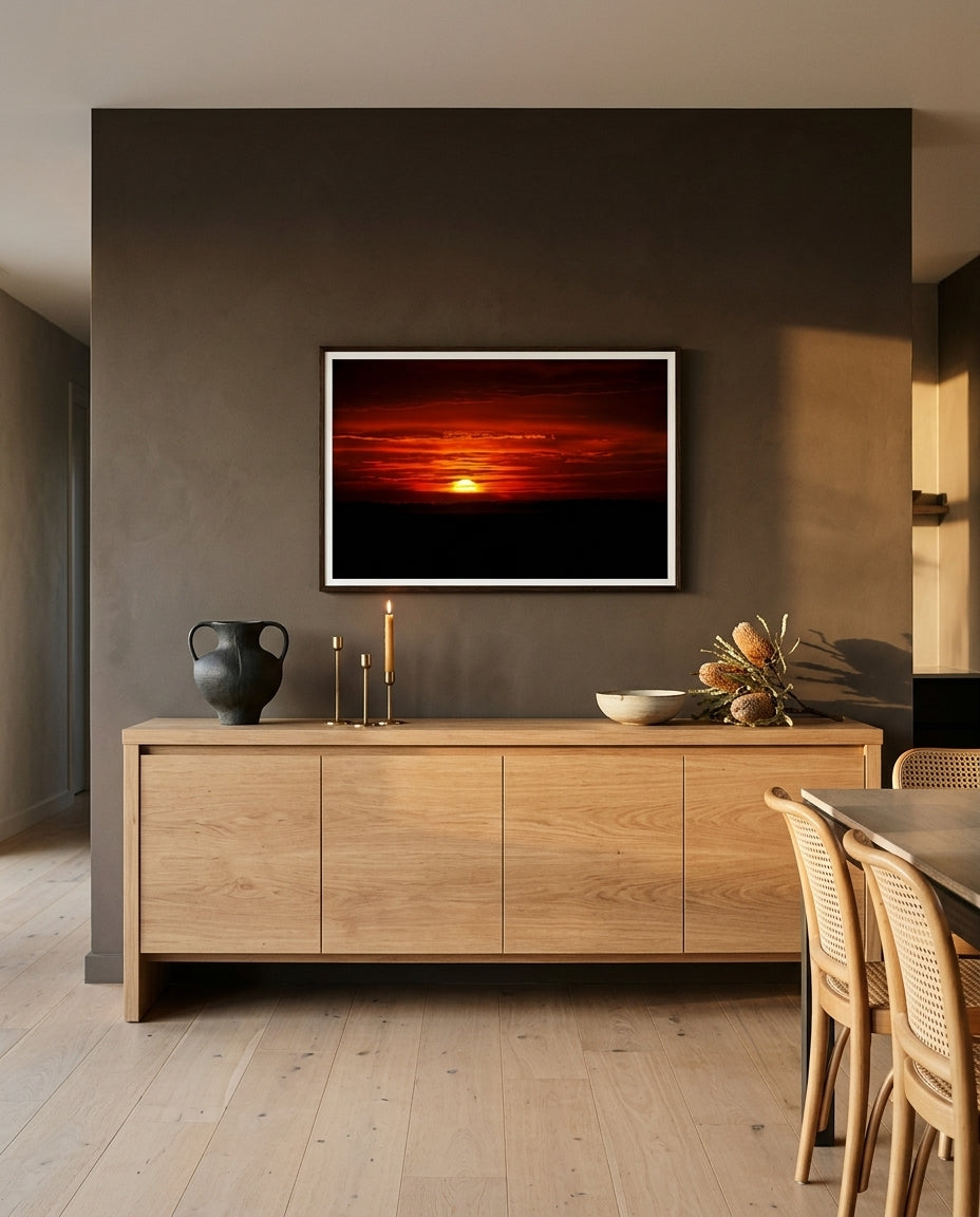 Sunrise Over Bass Strait – Flinders, framed above a long oak sideboard in an open-plan living-and-dining room — clear glass vessel, photography books, brass candleholder, ceramic bowl, dark wood frame. — Two Fat Bunnies.