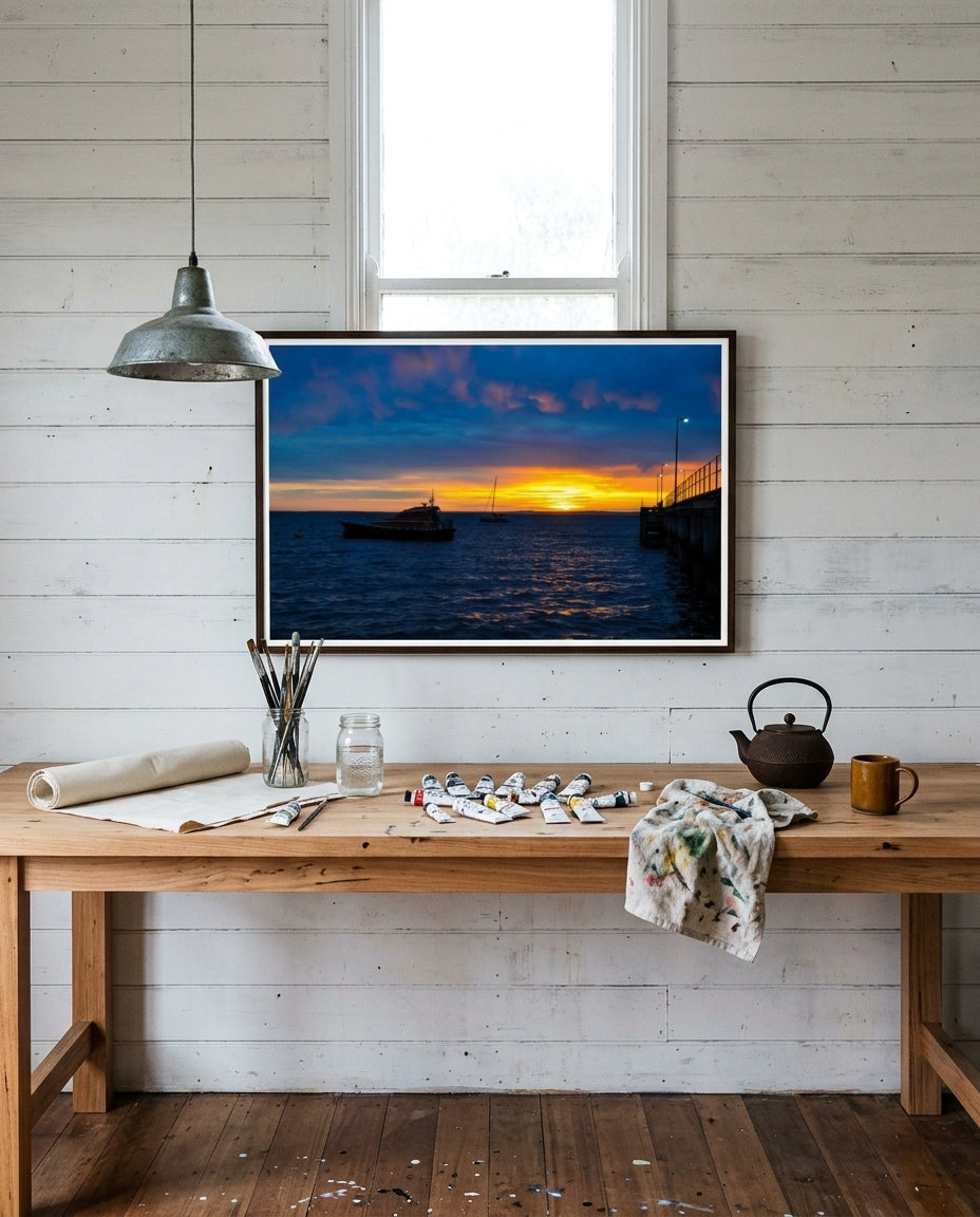 Sunrise Serenity at Flinders Pier, framed above a long pine workbench in a boatshed creative studio — exposed timber rafters, brass clamp lamp, sketchbooks, ceramic mug of brushes, dark wood frame. — Two Fat Bunnies.