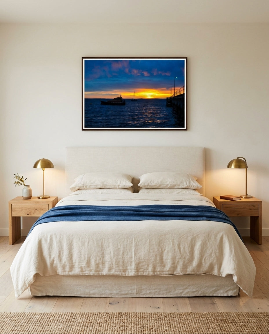 Sunrise Serenity at Flinders Pier, framed above a linen-headboarded bed in a Mornington master bedroom — cream linen, soft-blue wool throw, paper-shaded bedside lamps, oak side tables, dark wood frame. — Two Fat Bunnies.